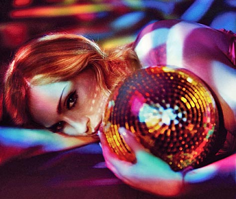 A person with red hair lying on their side, holding a disco ball against a colorful, patterned background of light.