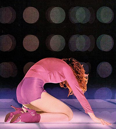 A woman in a pink outfit and high heels is kneeling on a glowing surface, with her back arched and head down, against a backdrop of softly blurred colorful lights.