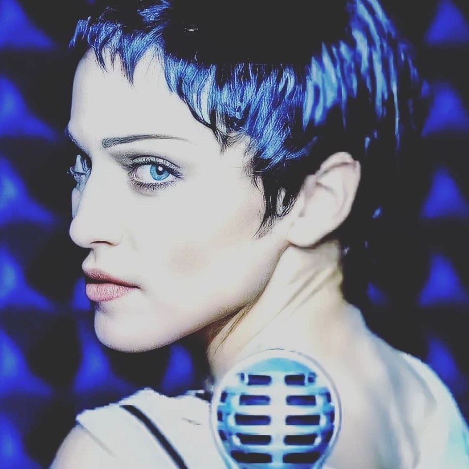 Profile view of a woman with short, blue-tinted hair and striking blue eyes, standing near a vintage microphone against a blue textured background.