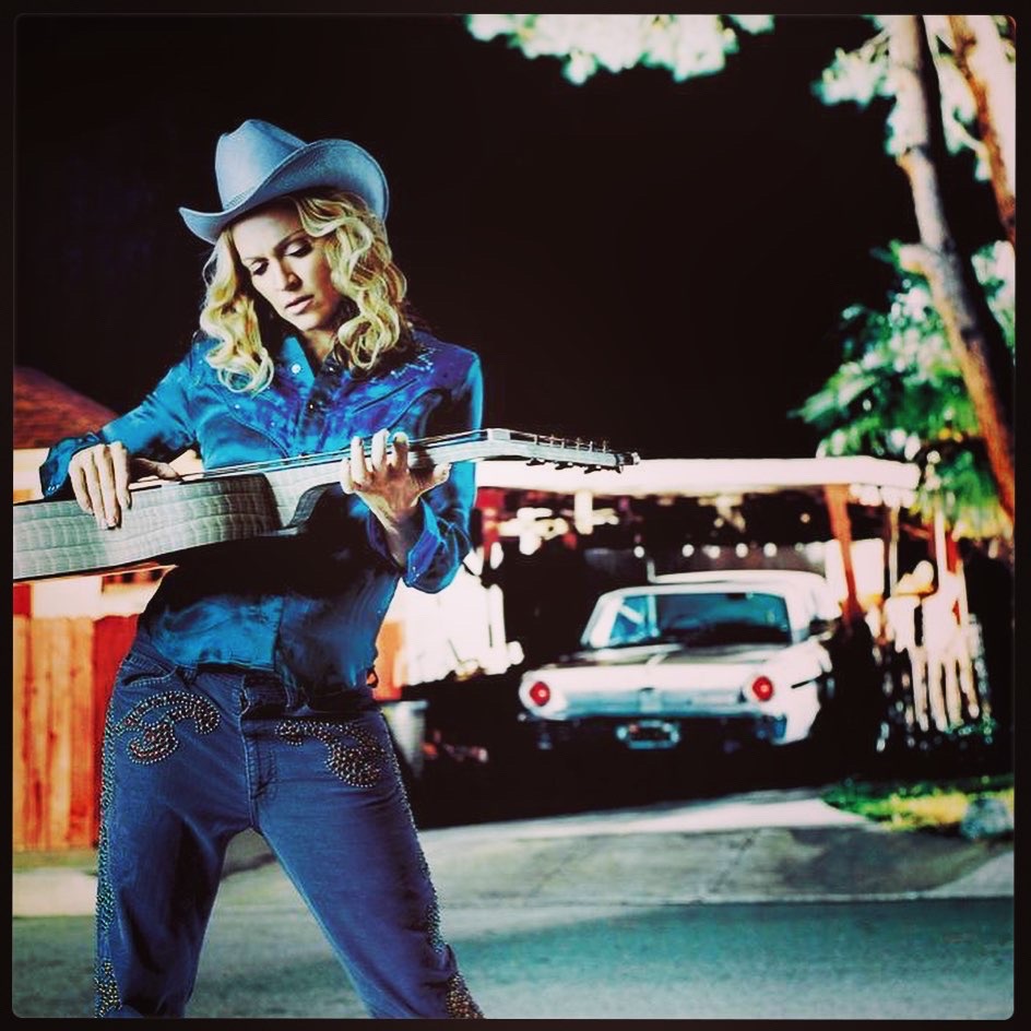 A woman wearing a cowboy hat and a blue shirt is playing a guitar outdoors at night, with a vintage car visible in the background.