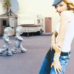 A woman in a white tank top and blue jeans stands with a playful pose, looking back over her shoulder. In the background, two figures in silver astronaut costumes walk by a trailer.