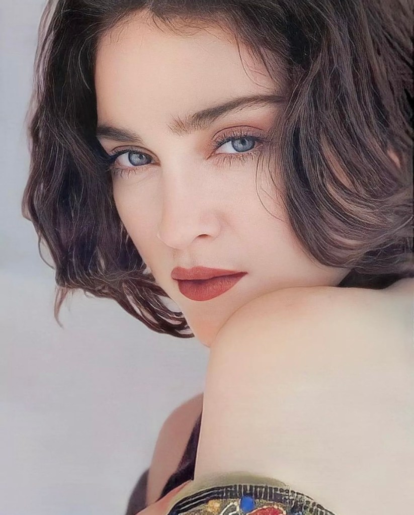 Close-up portrait of a woman with curly dark hair and striking blue eyes, wearing a deep red lip color, gazing intently at the camera.