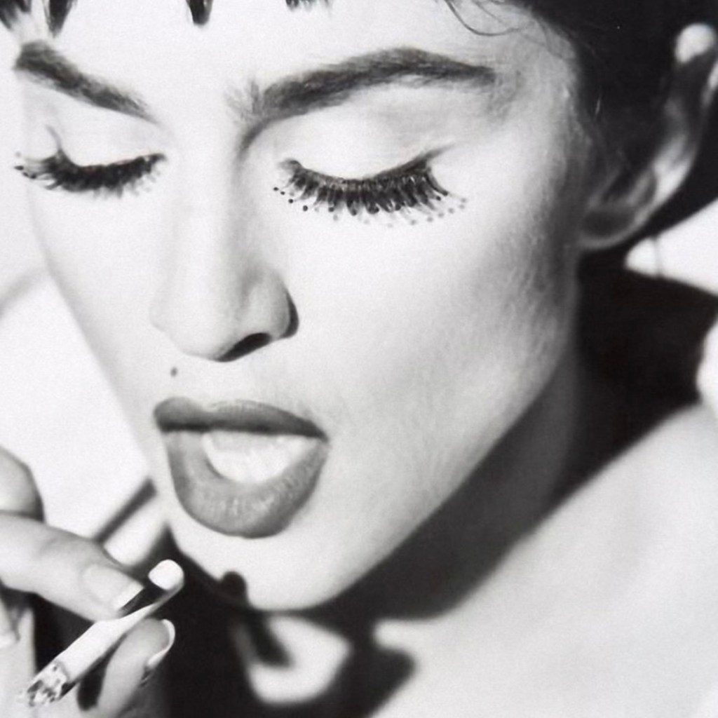 Black and white portrait of a woman with dramatic makeup, holding a cigarette near her lips, with long false eyelashes.