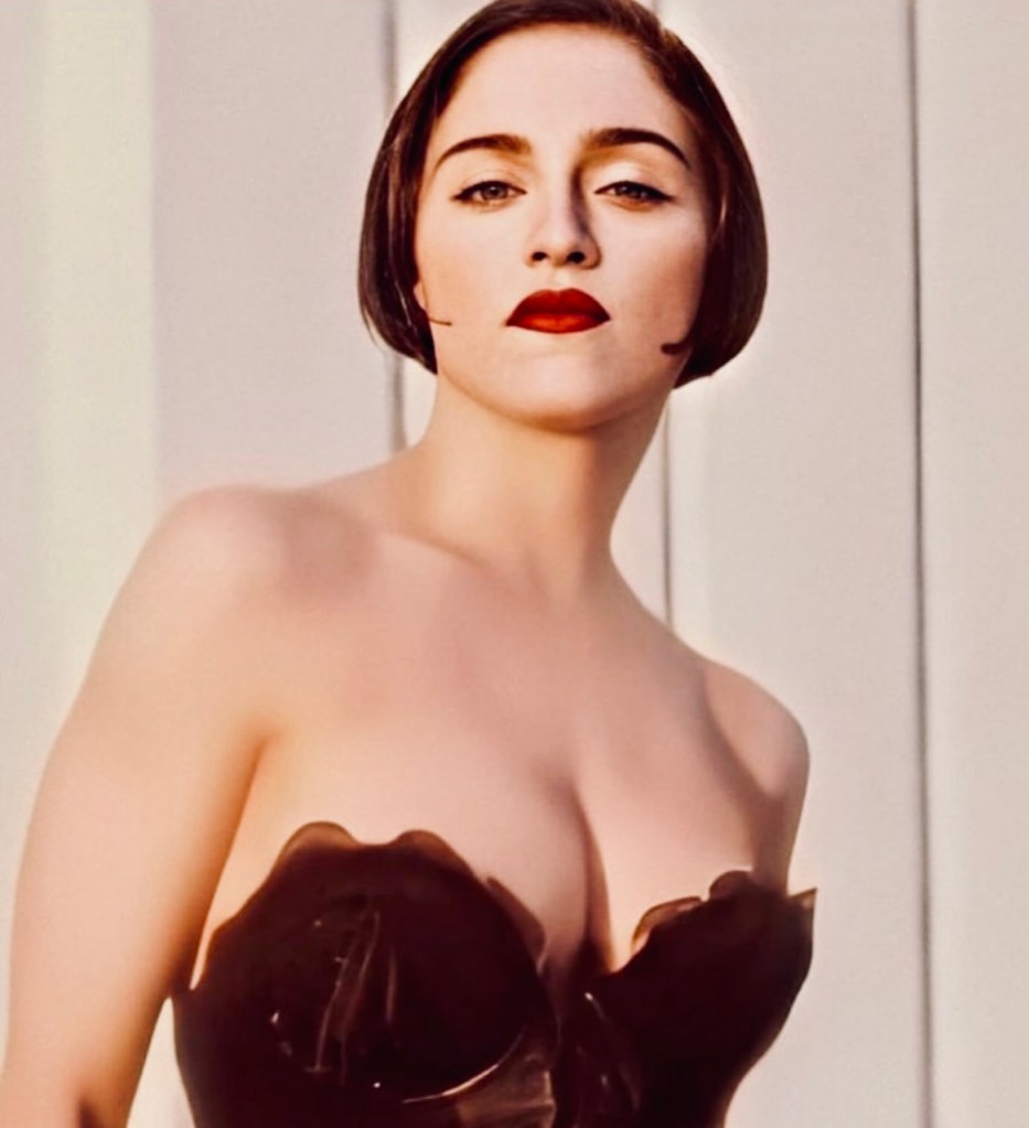A woman with short hair and red lipstick poses confidently in a strapless black top against a light background.