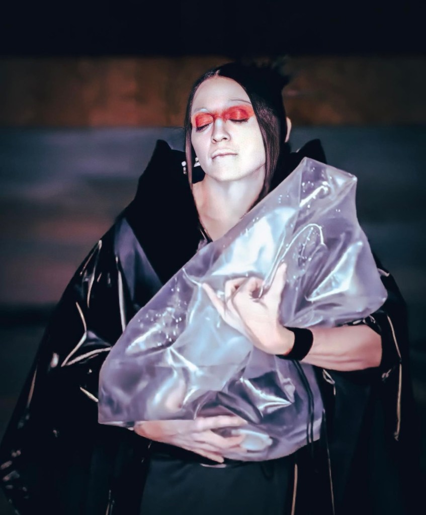 A person with closed eyes wearing dramatic makeup and a black outfit, holding a translucent plastic material close to their chest.