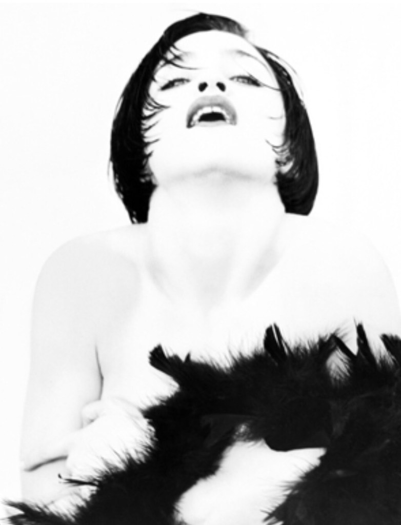 A black and white artistic portrait of a woman with short hair, looking upwards with a slightly open mouth, draped in a black feather boa.