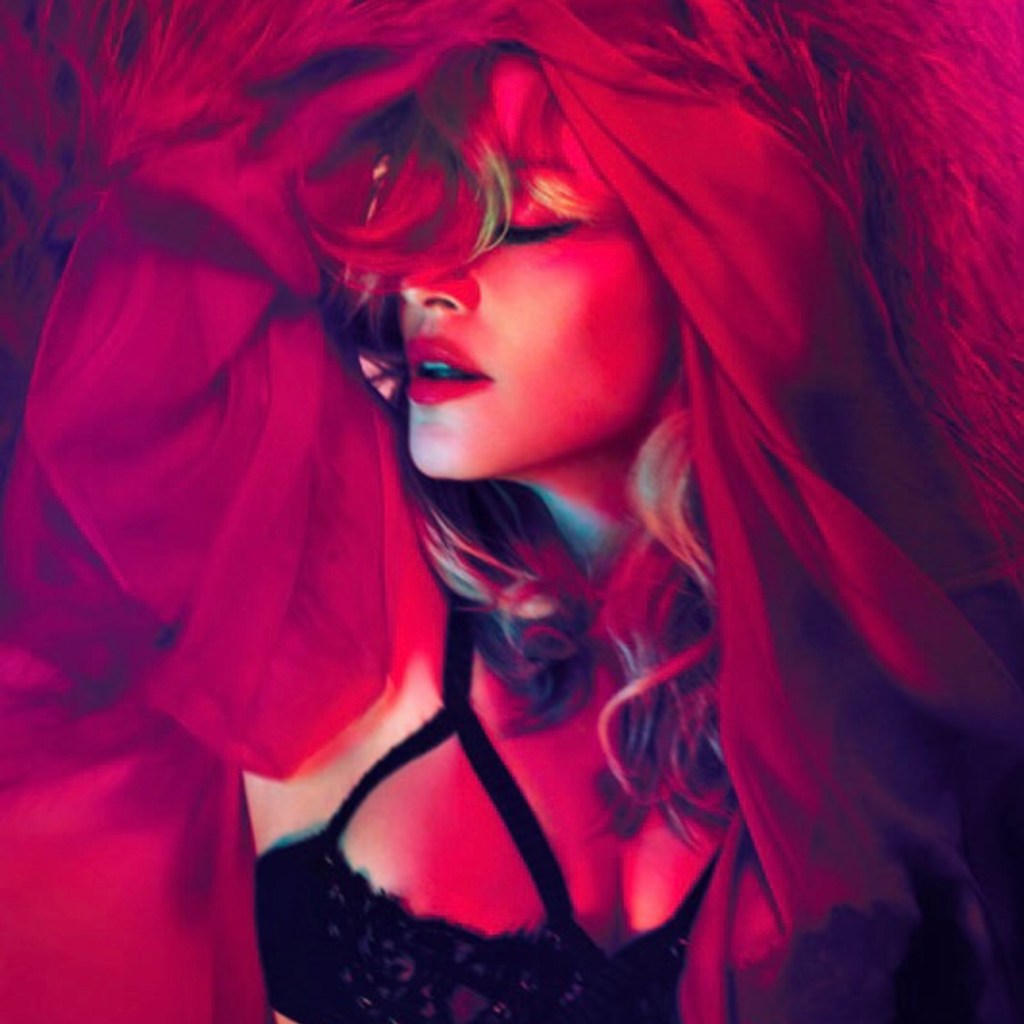 A close-up of a woman with wavy hair partially covered by a red veil, featuring dramatic lighting in shades of pink and red. She appears to be wearing a black lace outfit, with her eyes closed and a serene expression.