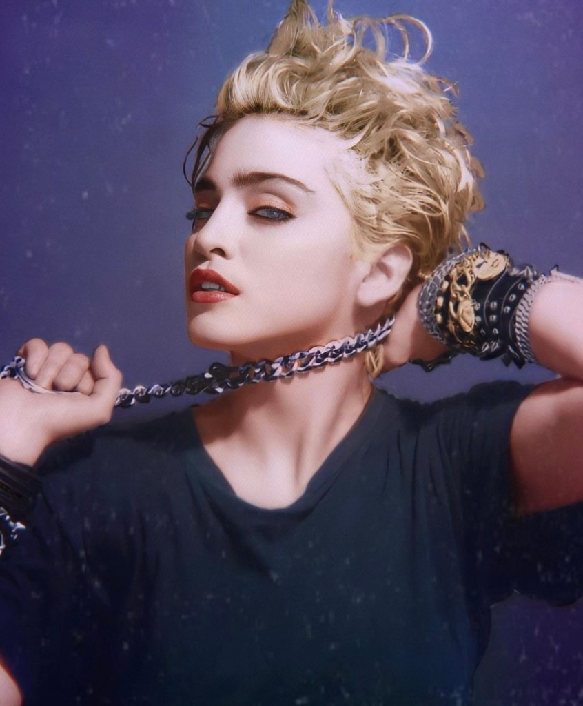 A blonde-haired individual with short, styled hair, wearing a black shirt and multiple bracelets, striking a pose while holding a chain.
