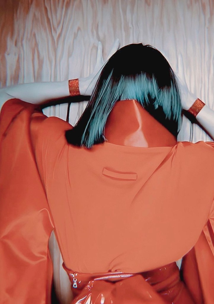 A person in a vibrant red outfit with glossy material, facing a wooden wall, with their hands resting on their head and hair styled in a sleek fashion.