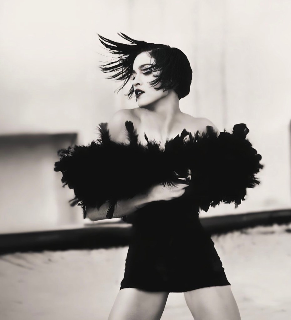 A woman in a black feathered garment poses dramatically with her hair blowing, captured in a black and white photograph.