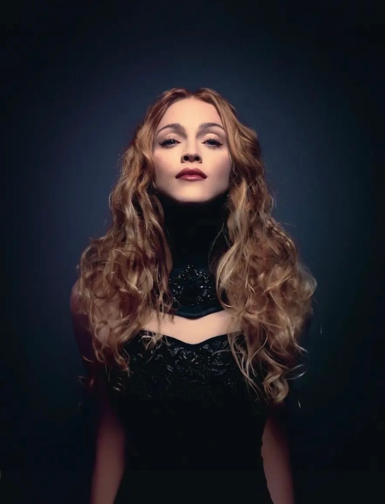 A woman with long, curly hair poses confidently in a dark backdrop, wearing an elegant black dress with a high neckline.