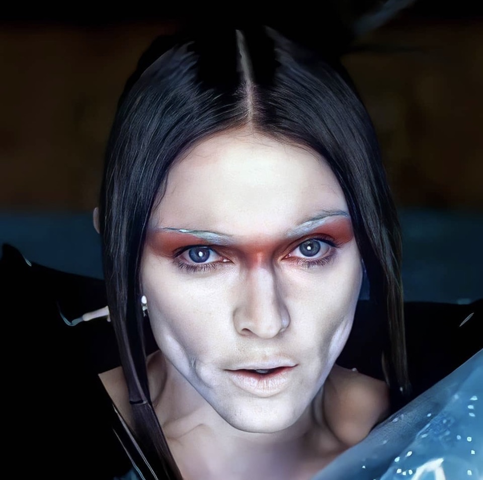 Close-up portrait of a person with striking facial makeup featuring bold eye shadow, pale skin, and visible facial contours, against a dark background.