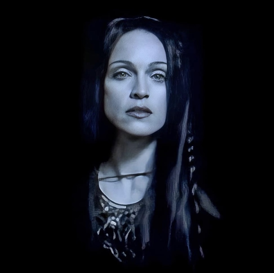 A portrait of a woman with long dark hair and striking features, set against a black background, with a cool blue tint.