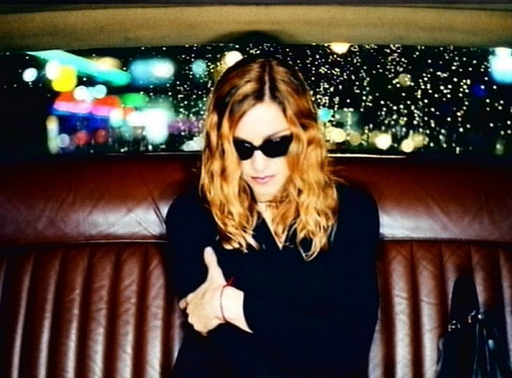 A woman with long, wavy, blonde hair wearing sunglasses, sitting in the back seat of a car, with a rainy cityscape illuminated by colorful lights visible through the window.