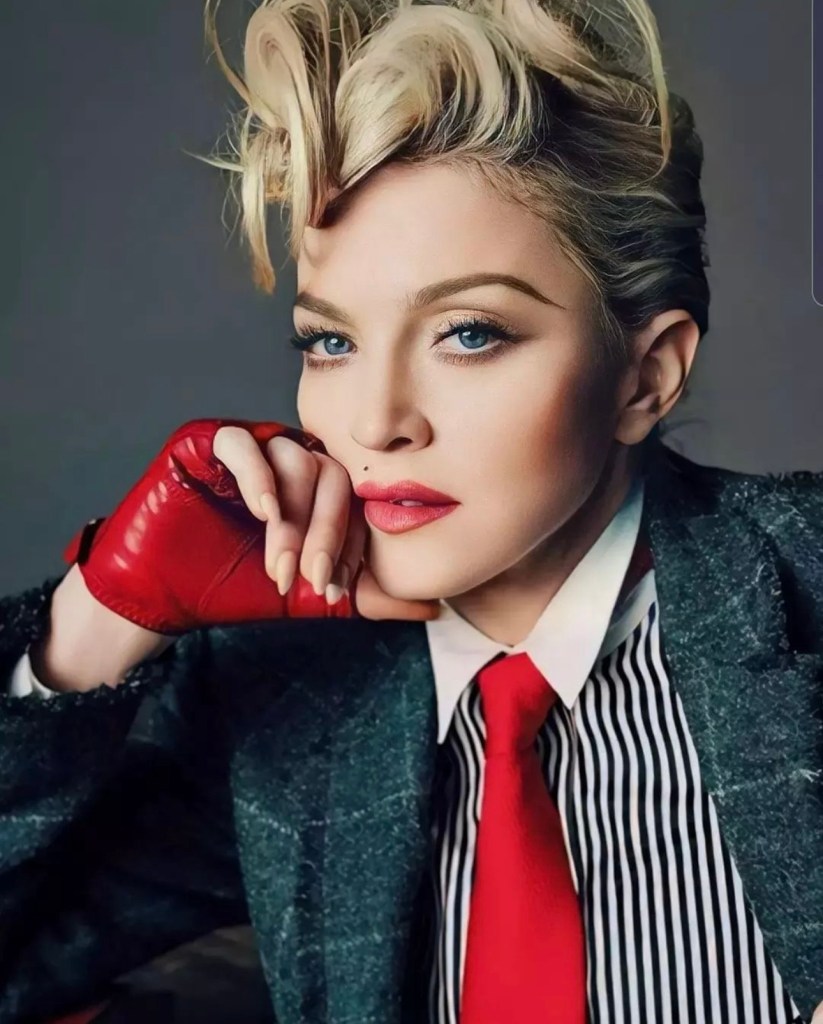A close-up portrait of a woman with styled blonde hair, wearing a striped shirt and a bold red tie, with a red leather glove resting on her chin.