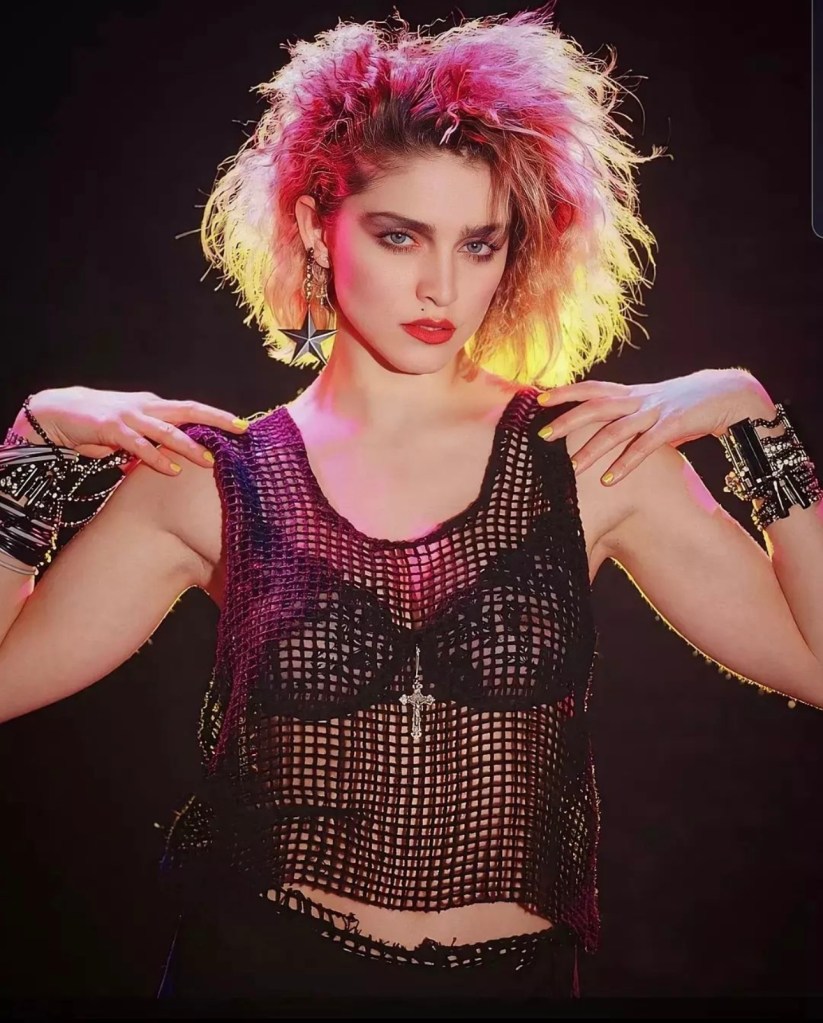 A model with curly hair styled in a vibrant 80s look, wearing a black fishnet top over a black bra and accessorized with bold earrings and bracelets, posing with a confident expression against a colorful background.