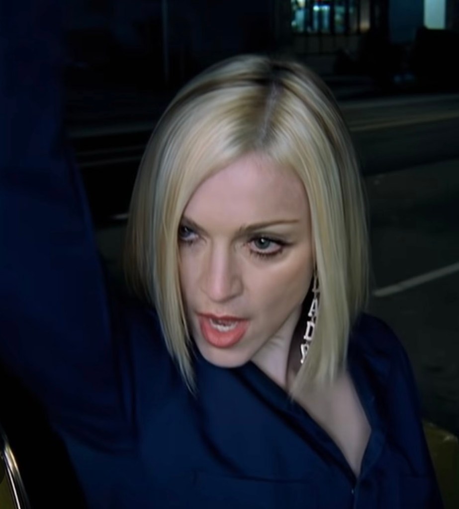 A close-up of a blonde woman with a stylish bob haircut, wearing a dark blue shirt and large earrings, looking intensely while raising one arm.
