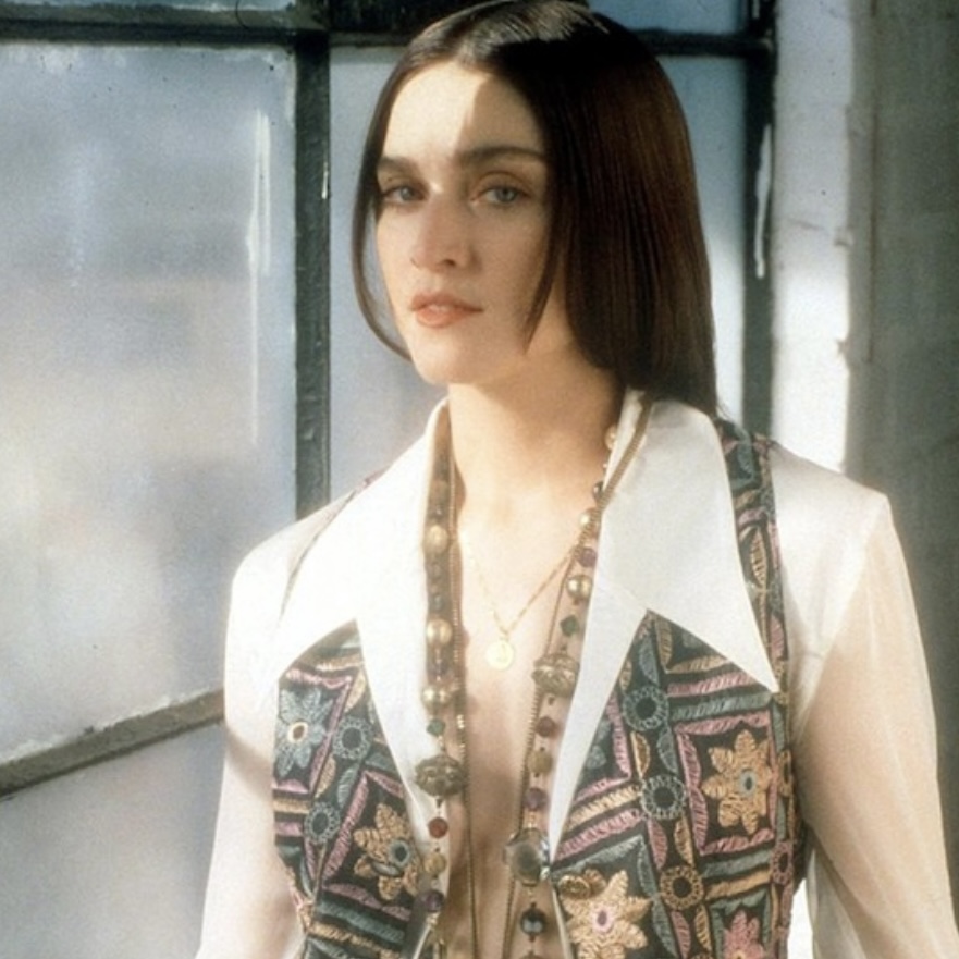 A woman with long hair stands in front of a window, wearing a vintage patterned vest over a light blouse, with layered necklaces and a serious expression.
