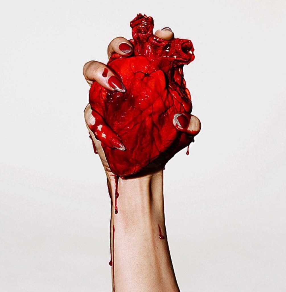 A hand gripping a realistic red heart, with blood dripping, set against a white background.