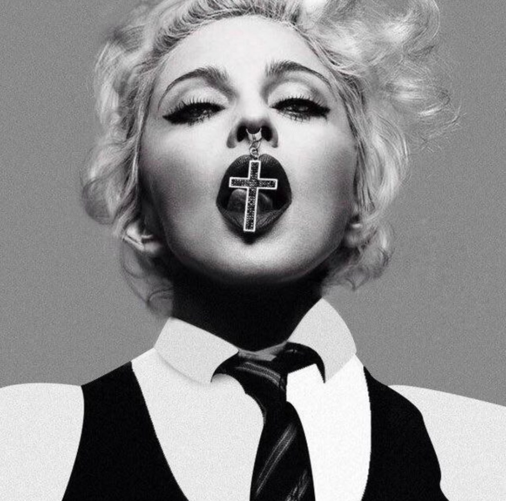Close-up black and white portrait of a woman with short, curly blonde hair, wearing a tie and collared shirt. She has a cross pendant hanging from her mouth, with bold eye makeup and a striking expression.
