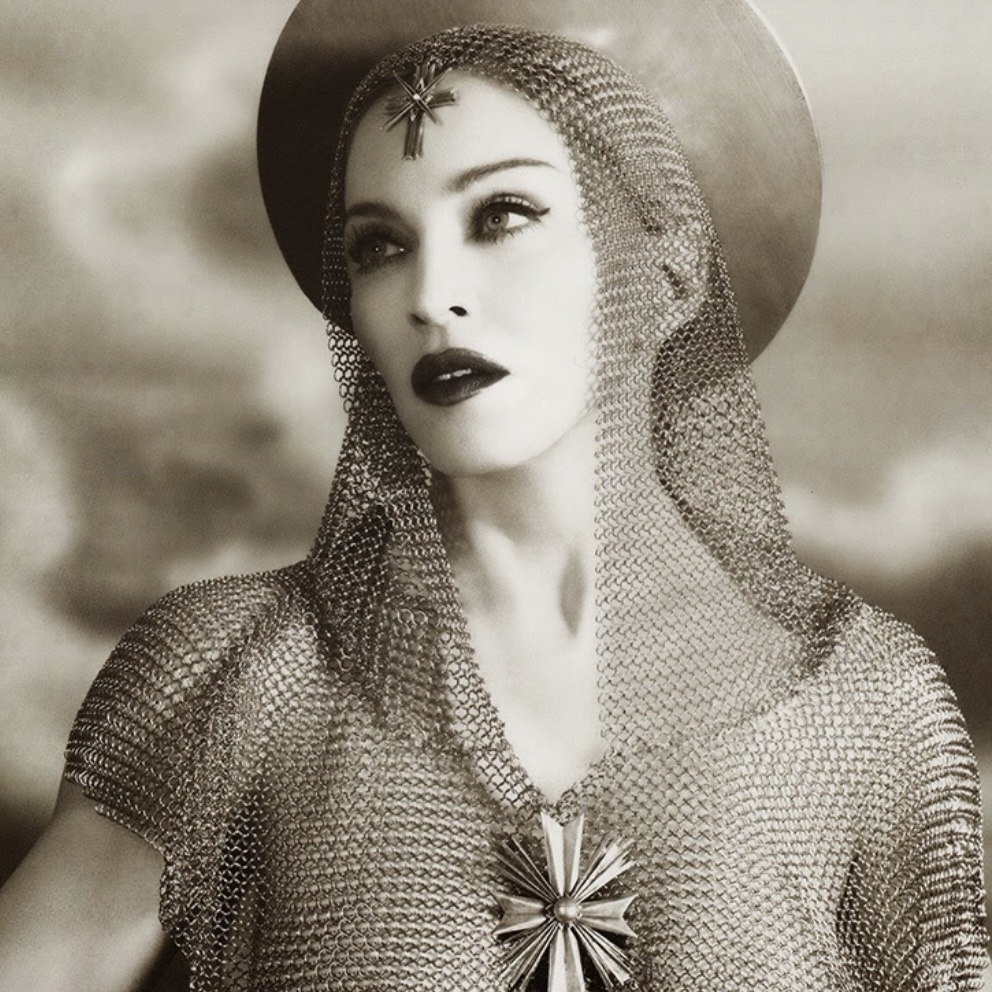 A black-and-white portrait of a woman wearing a chain mail headdress and a jeweled cross, gazing upward with a serene expression.