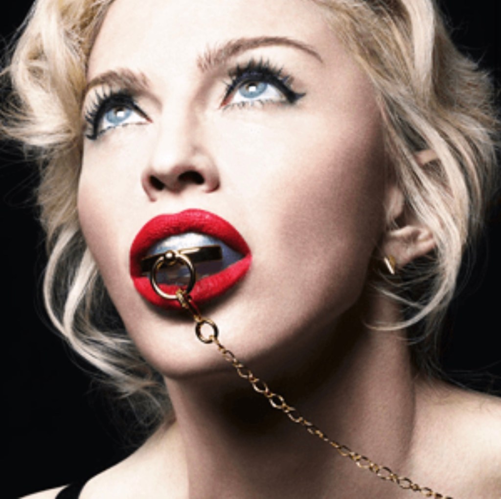 A close-up portrait of a woman with platinum blonde hair, striking blue eyes, and bold red lipstick. She is looking upwards with a silver object in her mouth attached to a gold chain.