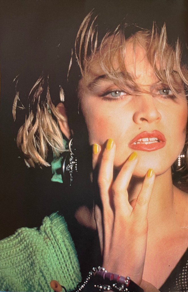 Close-up portrait of a stylish woman with short, wavy hair and bold makeup, wearing bright yellow nail polish, set against a dark background.