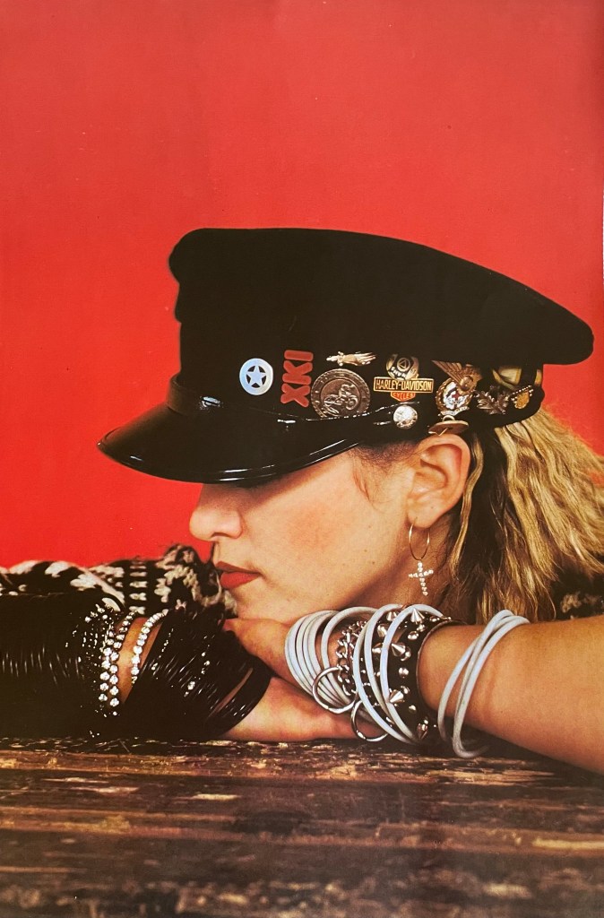 Profile view of a woman wearing a black cap adorned with various badges and jewelry, resting her chin on her crossed arms, with a red background.