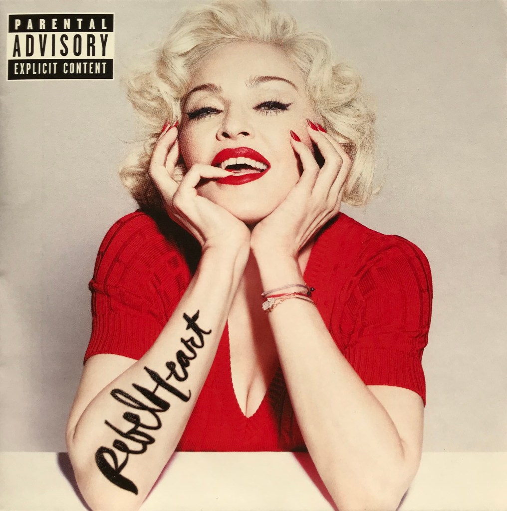 A person with blonde, curly hair smiles and poses with hands on their face, wearing a red top. The arm displays the tattoo 'Rebel Heart', and a parental advisory label is present in the top corner.