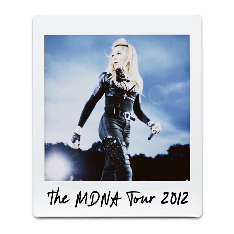 A performer dressed in a black outfit with gloves and a cross necklace stands on stage during The MDNA Tour in 2012, with a spotlight illuminating her.