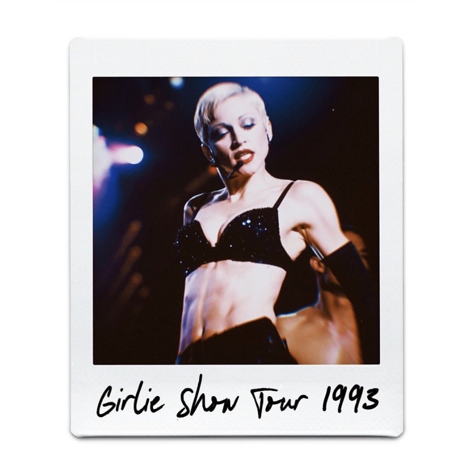 A performer dressed in a sparkling black bra and gloves on stage during a concert, with dramatic lighting in the background; labeled 'Girlie Show Tour 1993'.