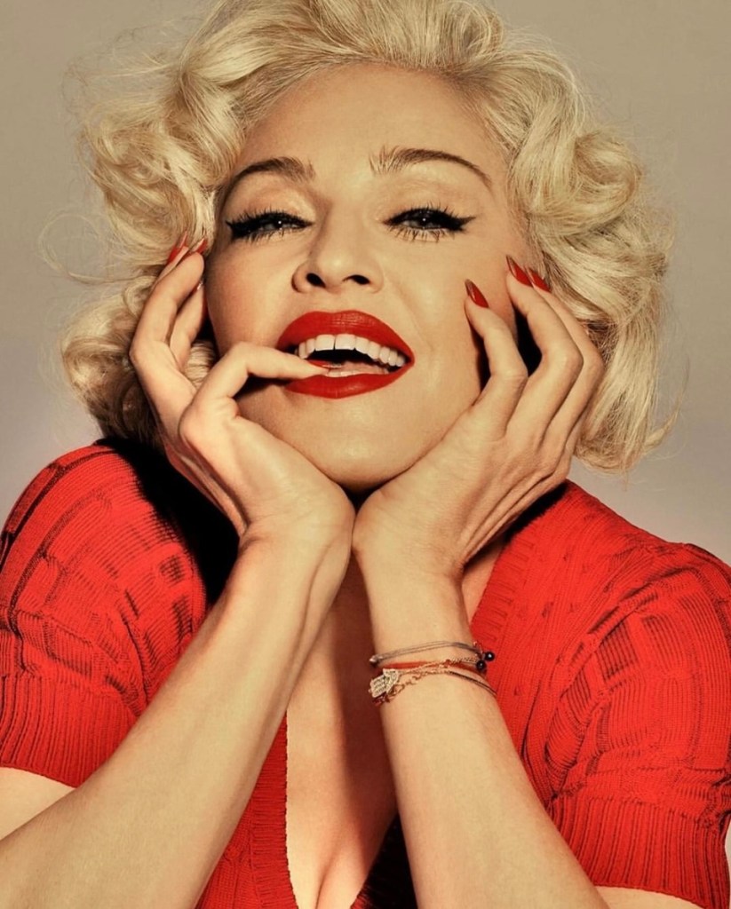 A woman with blonde, curly hair smiles warmly, showcasing bright red lipstick. She has a playful expression, resting her hands on her face, and is wearing a red top.