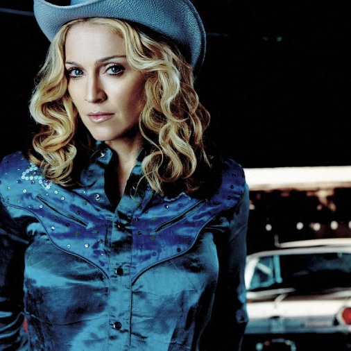 A woman with long, wavy blonde hair wearing a blue cowboy shirt and a light blue cowboy hat, posing confidently in front of a vintage car.