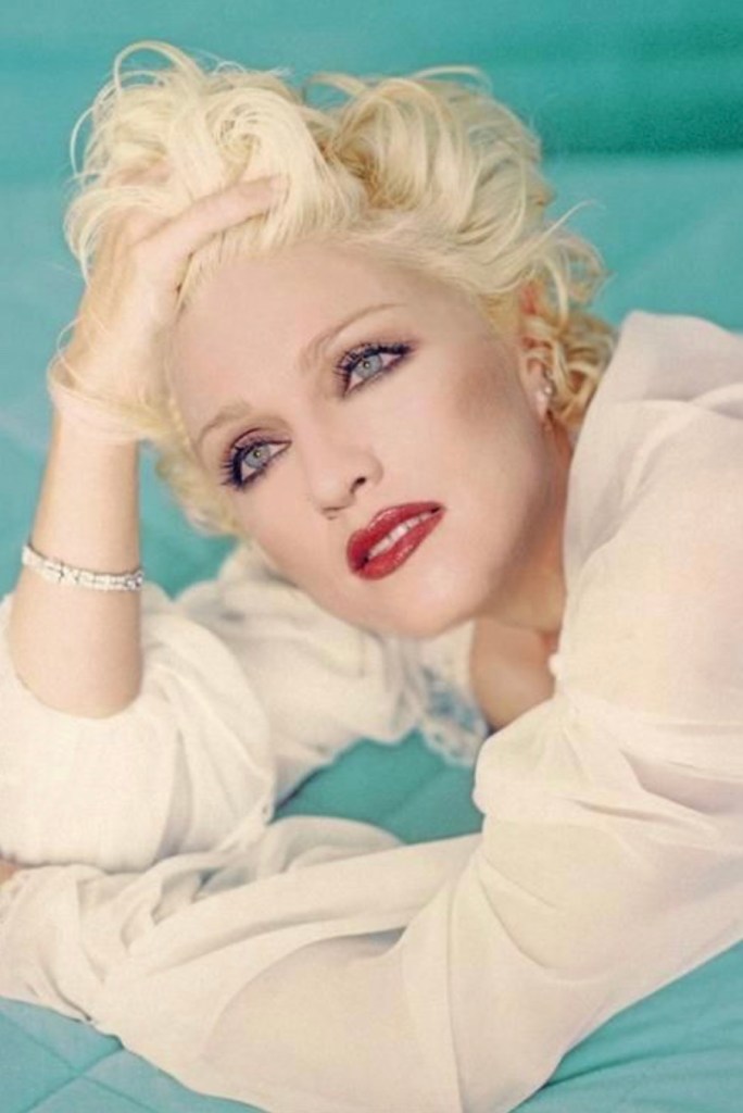A close-up portrait of a woman with short, styled blonde hair, reclining on a turquoise background. She has striking blue eyes, bold red lipstick, and is wearing a sheer white garment. Her hand is gently placed on her head, conveying a contemplative expression.