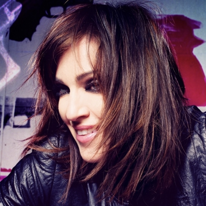 A close-up portrait of a woman with long, flowing hair, wearing a black leather jacket, smiling against a colorful graffiti background.
