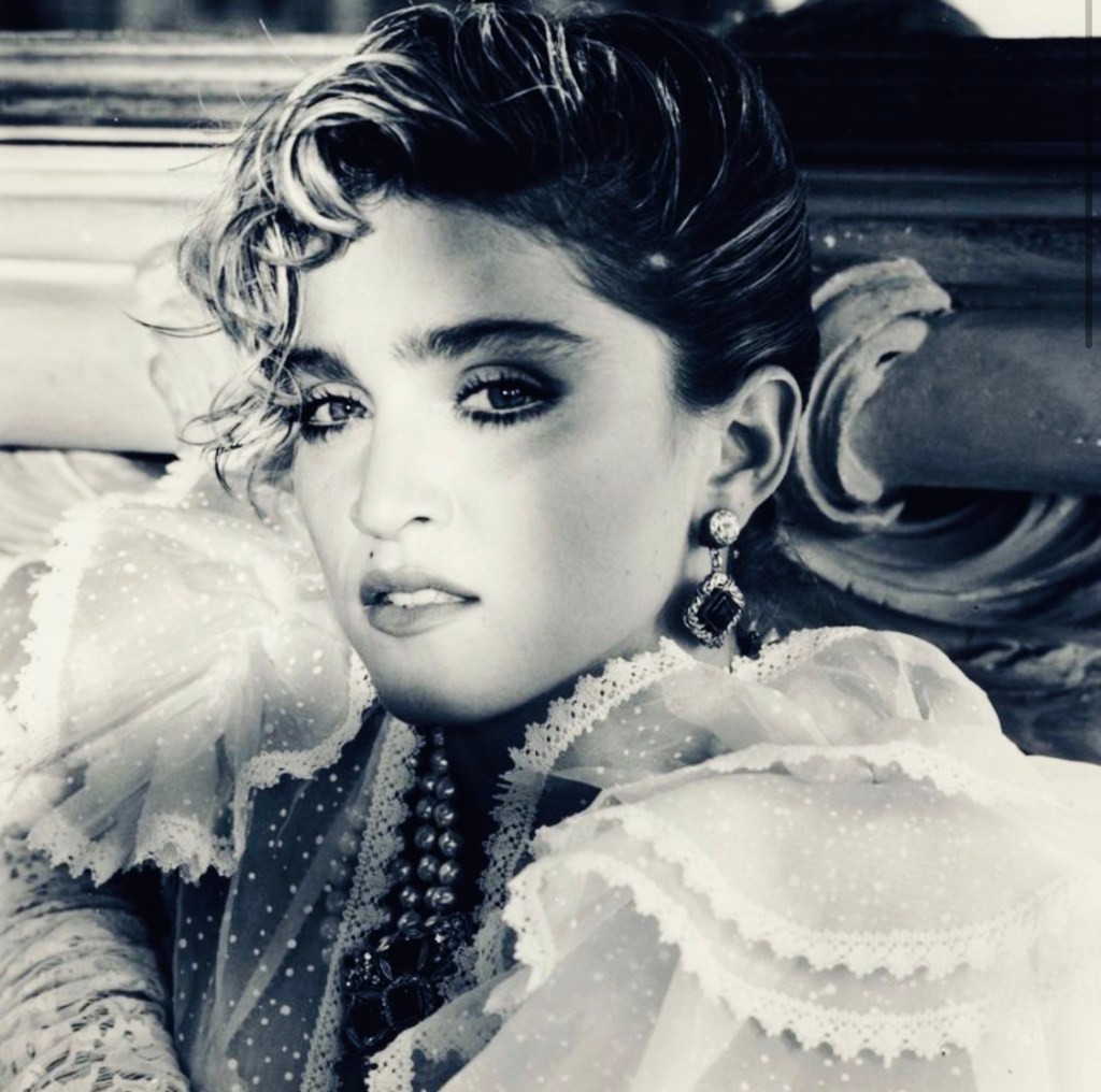 Black and white portrait of a woman with short, wavy hair and bold makeup, wearing a ruffled lace blouse and large earrings, gazing intently at the camera.