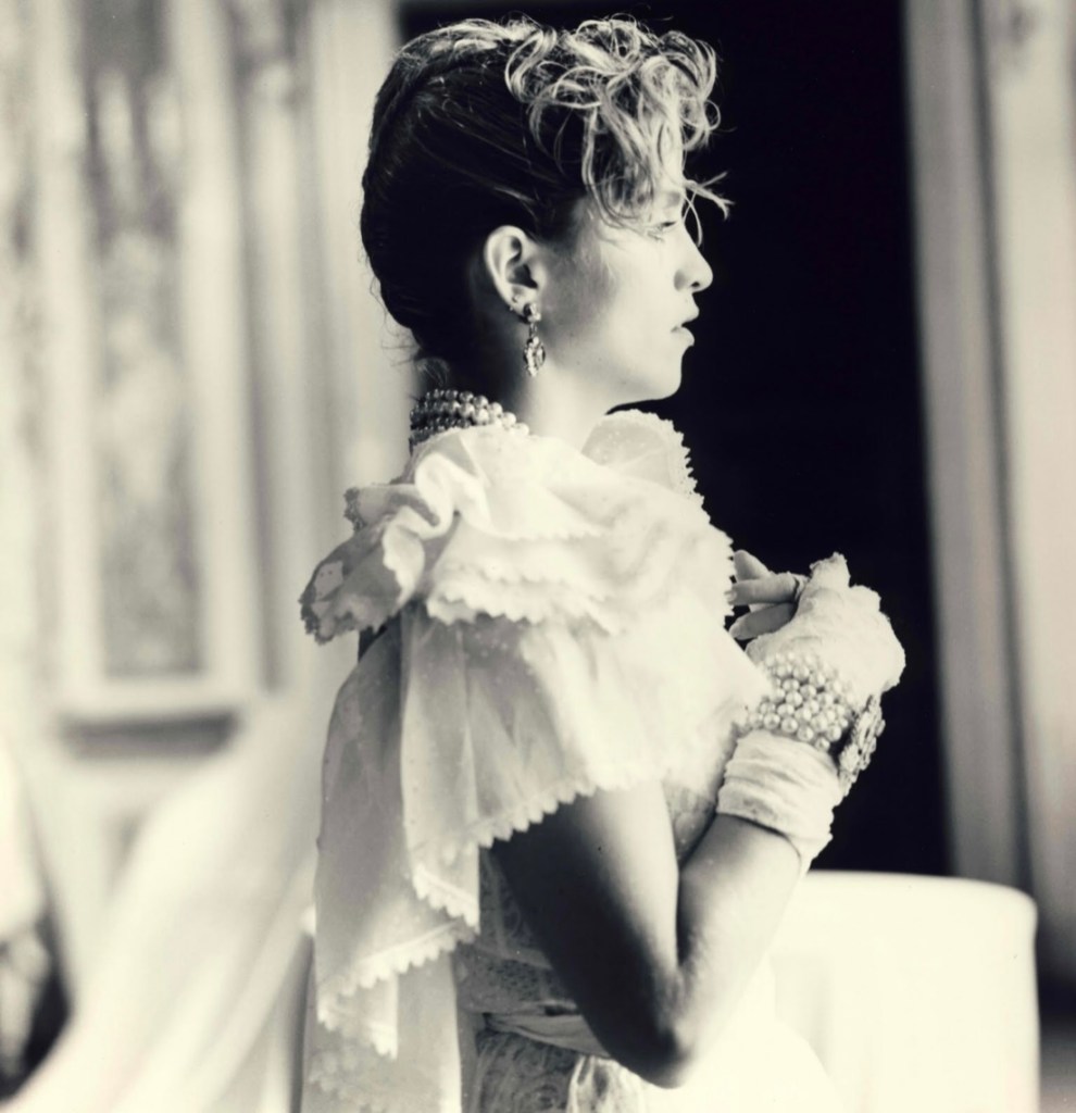 A side profile of a woman wearing a vintage white dress with ruffled details, adorned with pearls and gloves, set against a softly blurred background.
