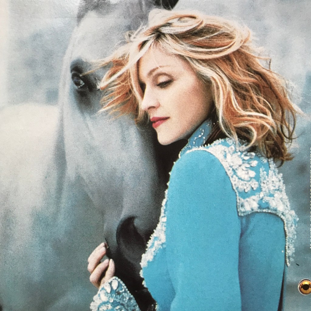 A woman with wavy hair, wearing a blue embellished jacket, softly leans against a gray horse, creating an intimate and serene atmosphere.