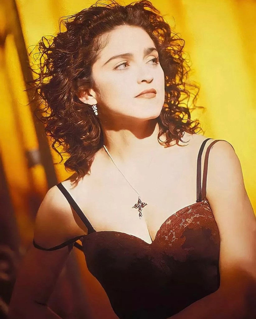A woman with curly hair wearing a dark lace dress, posing against a warm yellow background.