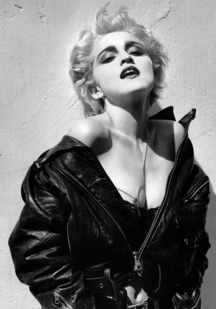 Black and white portrait of a woman in a leather jacket, striking a confident pose with her head tilted slightly back and lips pursed. The background is a plain wall, enhancing the focus on her expression and style.