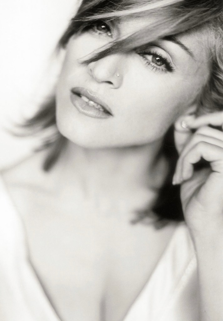A close-up black and white portrait of a woman with short hair, showcasing a thoughtful expression. She has a nose piercing and is partly touching her face.