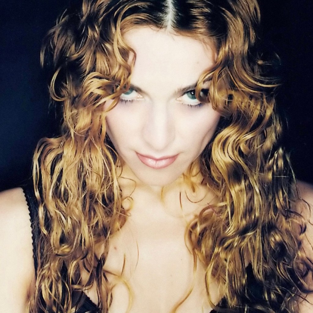 A close-up portrait of a woman with curly blonde hair, staring intensely at the camera against a dark background.
