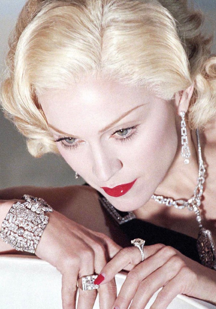 Close-up portrait of a woman with blonde hair, wearing elegant jewelry and red lipstick, looking thoughtfully down.