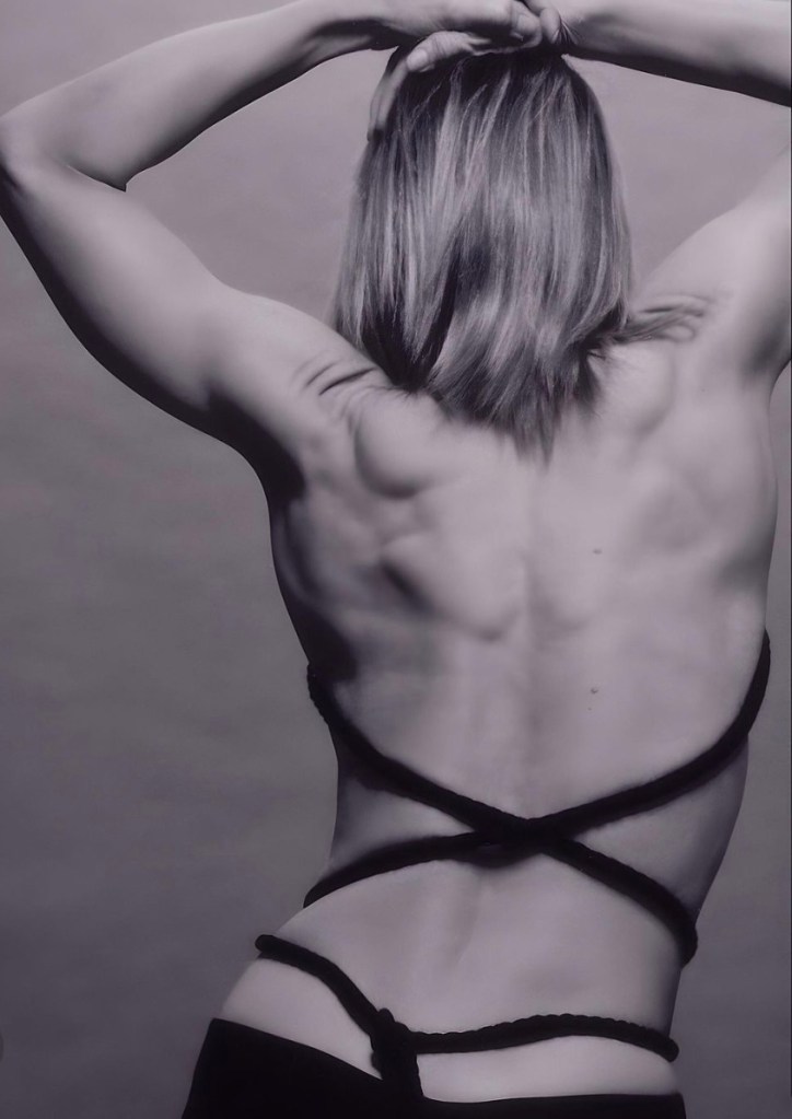 Black and white image of a woman from the back, showcasing toned muscles and a hairstyle that falls just above the shoulders. She is wearing a top with intricate straps crisscrossing over her back.