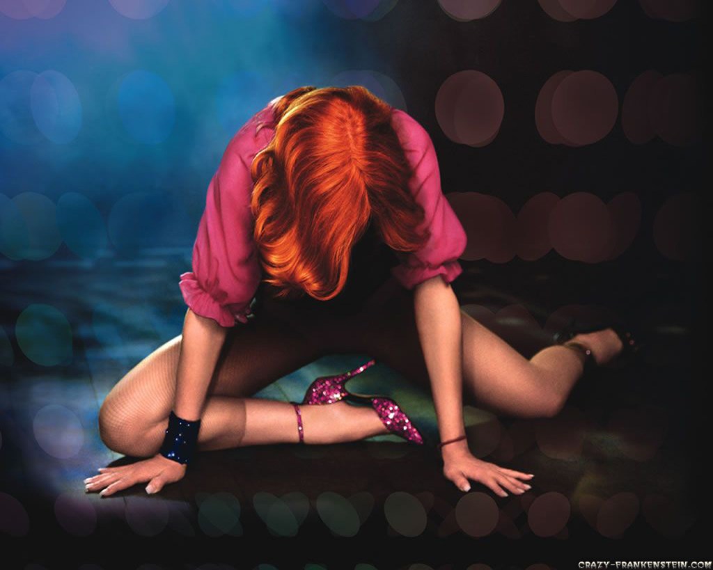 A person with vibrant red hair, wearing a pink blouse and black fishnet stockings, is positioned on the floor in a dramatic pose, surrounded by a softly blurred background.