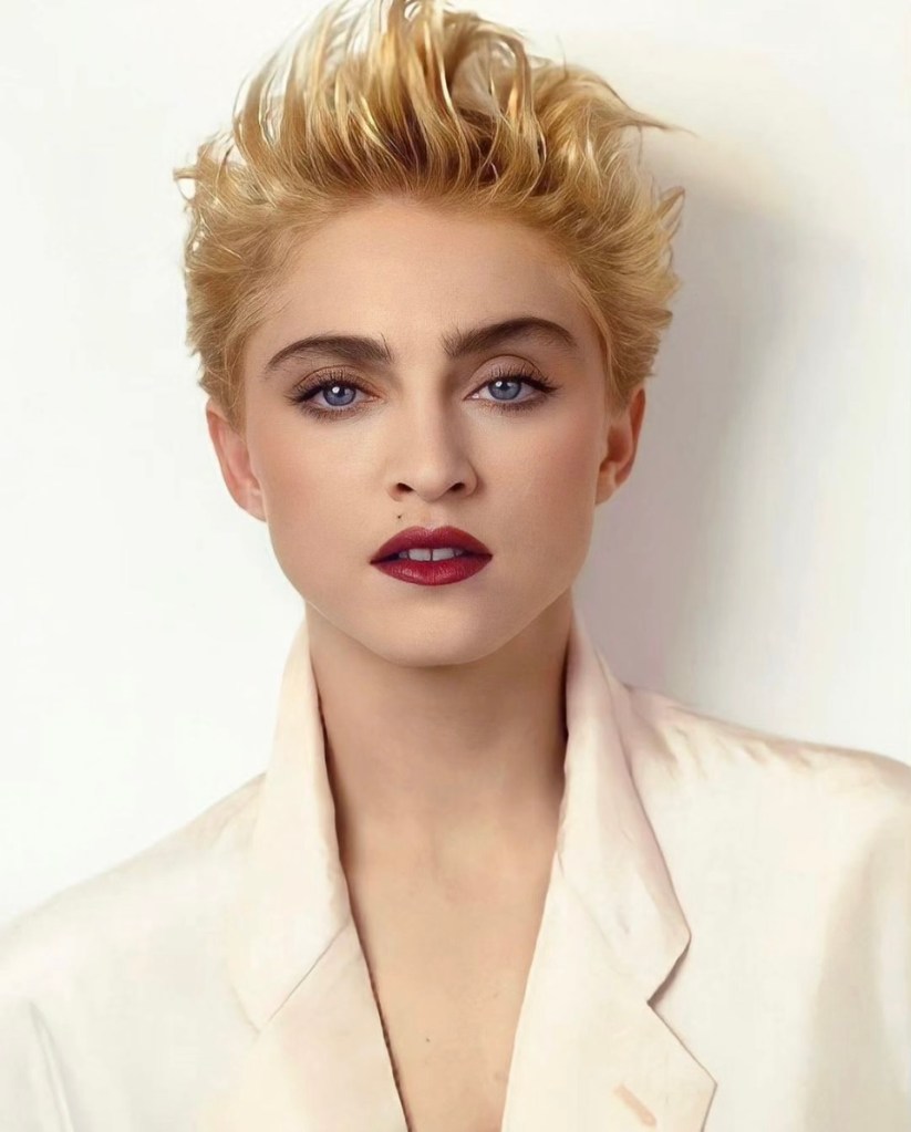 Close-up portrait of a person with short blonde hair, blue eyes, and bold red lipstick, wearing a light-colored blazer