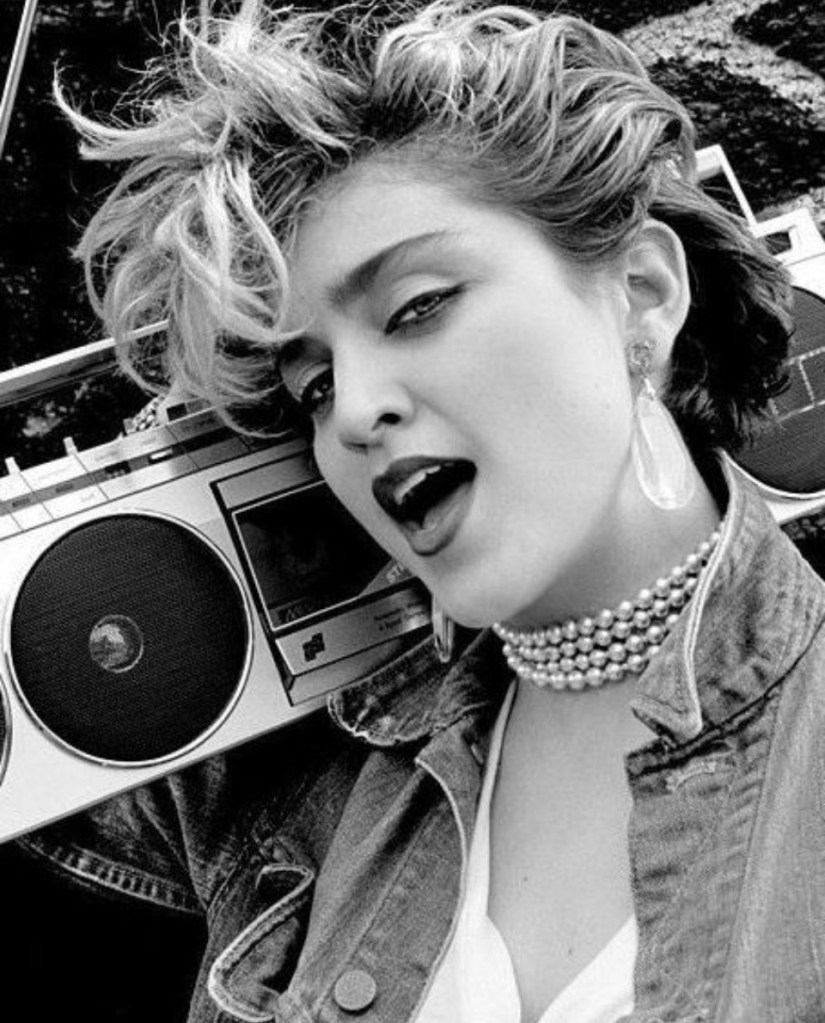 A black and white portrait of a woman with stylish, short, wavy hair, wearing a denim jacket and pearl accessories, singing passionately while leaning against a boombox.