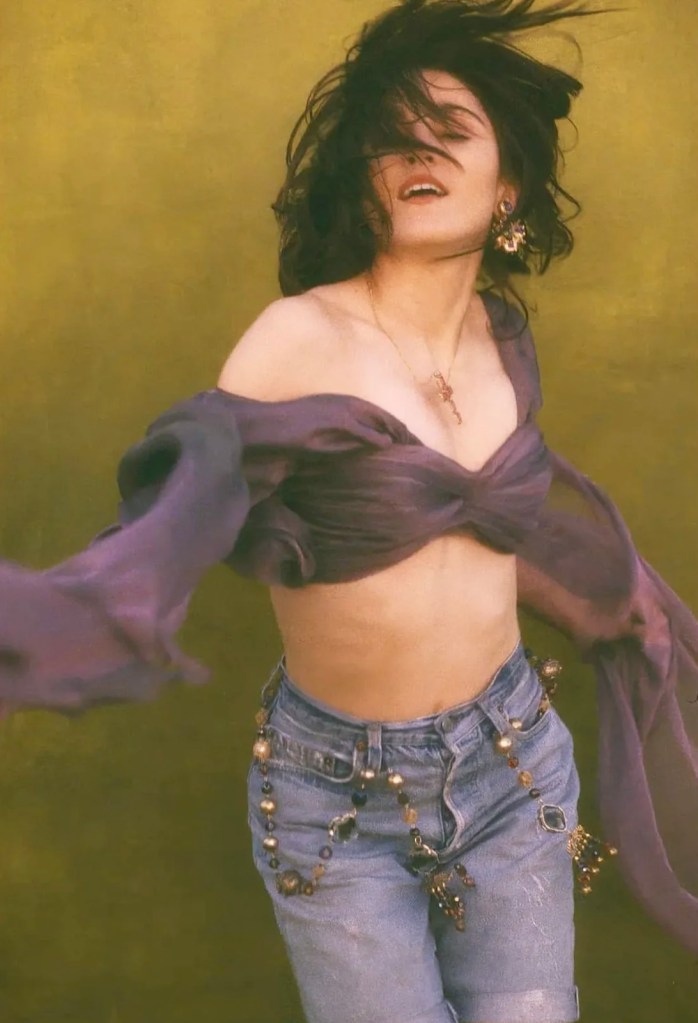 A person with flowing hair is dancing in front of a textured green background, wearing a purple top and high-waisted denim jeans adorned with decorative beads.