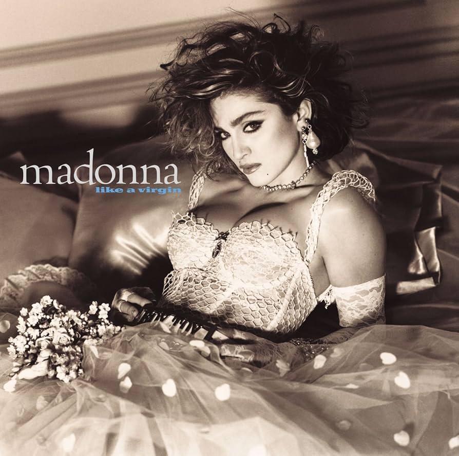 Black and white image of a woman styled in vintage fashion, lying on a bed with a bouquet of flowers, wearing a lace and polka dot outfit.