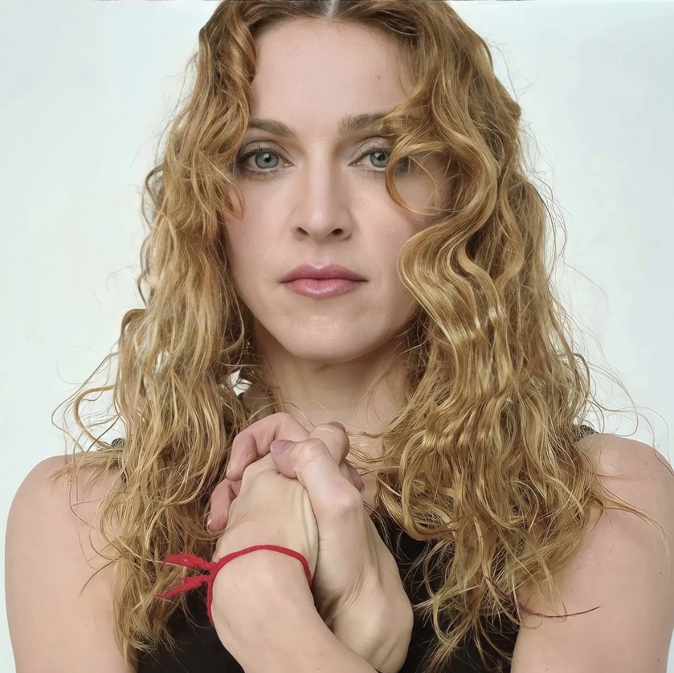 Portrait of a woman with wavy, shoulder-length blonde hair, holding her hands together in front of her chest. She has a serious expression, with soft makeup, and is wearing a black top. A red string bracelet is visible on her wrist.